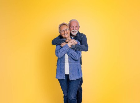 Foto de Retrato de un hombre mayor barbudo amoroso romántico abrazando a la esposa por detrás y mirando a la cámara mientras posan juntos sobre un fondo amarillo - Imagen libre de derechos
