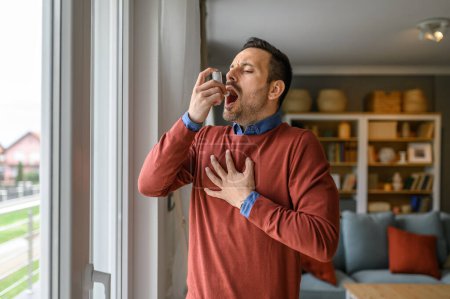 Foto de Joven sofocado que sufre dolor en el pecho inhalando a través de la bomba de asma mientras está en casa - Imagen libre de derechos