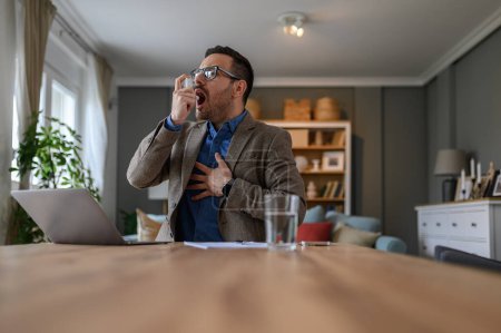Foto de Profesional masculino tocando el pecho con dolor e inhalando a través del inhalador de asma en el escritorio en la oficina en casa - Imagen libre de derechos