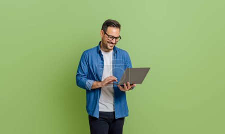 Photo for Smiling young businessman analyzing data over laptop while standing isolated on green background - Royalty Free Image