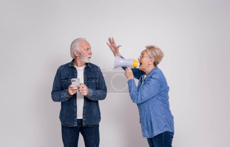 Shocked senior husband with smart phone looking at wife screaming over megaphone on white background