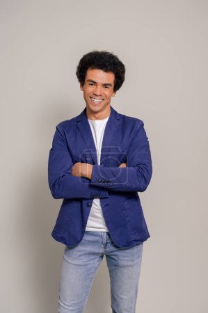 Glücklicher junger Geschäftsmann mit Afro-Haaren und verschränkten Armen posiert selbstbewusst vor weißem Hintergrund