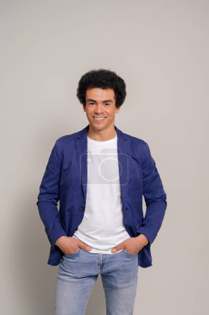 Portrait of happy male professional with afro hair and hands in pockets posing over white background