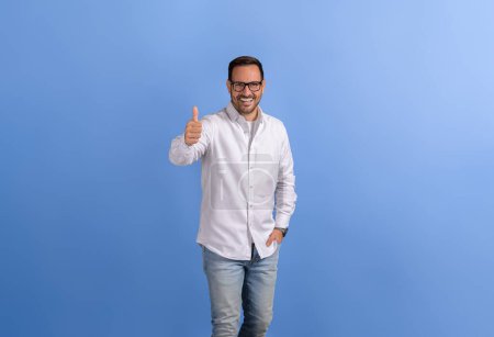 Porträt eines positiven männlichen Profis mit der Hand in der Tasche, die das Zeichen Daumen hoch auf blauem Hintergrund zeigt