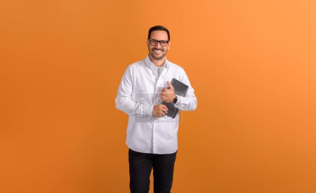 Successful young male freelancer holding digital tablet and smiling at camera on orange background