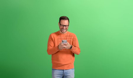 Retrato de empresario guapo sonriendo y mensajería en línea a través del teléfono inteligente sobre fondo verde