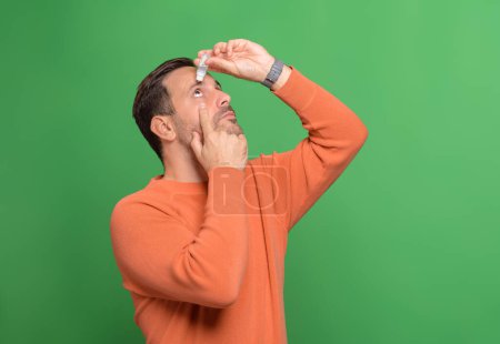 Jeune homme goutte lubrifiant pour les yeux pour traiter les yeux secs ou allergie tout en se tenant debout sur fond vert