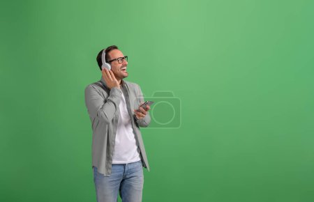 Hombre guapo con teléfono móvil escuchando música sobre auriculares y riendo sobre fondo verde