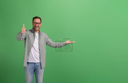 Verkäufer lacht und demonstriert Produkt auf Handfläche und zeigt Daumen hoch Zeichen auf grünem Hintergrund