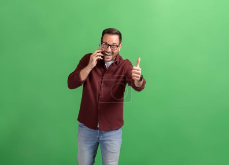 Successful young businessman talking on smart phone and showing thumbs up sign on green background