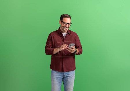Joven empresario guapo en gafas televisando a través del teléfono inteligente sobre fondo verde