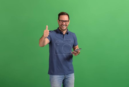 Joven empresario positivo con teléfono inteligente que muestra el signo de pulgares hacia arriba mientras posa sobre fondo verde
