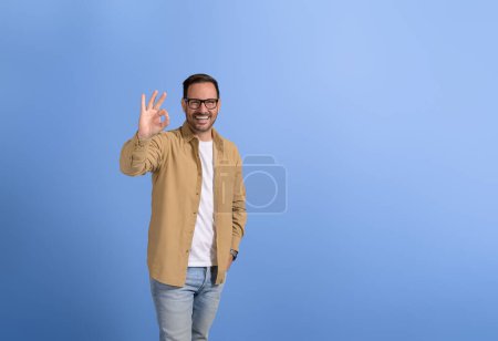 Porträt eines gut aussehenden Verkäufers mit lächelnder Hand in der Tasche und OK-Zeichen auf blauem Hintergrund