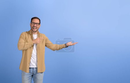 Jungunternehmer lacht und wirbt auf leerer Handfläche für neues Produkt, während er auf blauem Hintergrund steht