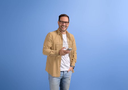 Junger glücklicher Geschäftsmann mit Hand in Tasche nutzt soziale Medien auf Smartphone vor blauem Hintergrund