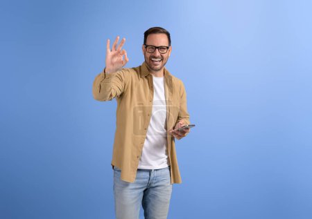 Gut gelaunter Unternehmer hält Smartphone in der Hand und gestikuliert mit OK-Zeichen auf blauem Hintergrund