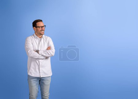Happy businessman with arms crossed contemplating and looking away while standing on blue background