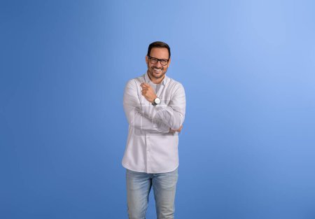 Handsome freelancer with arm crossed and in glasses smiling at camera and posing on blue background
