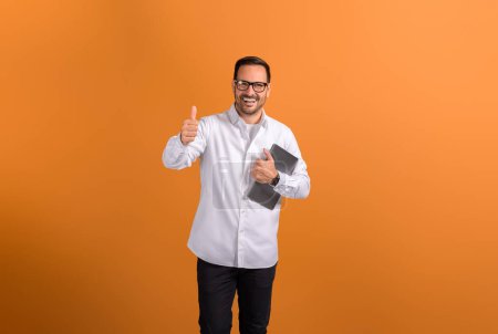 Erfolgreicher männlicher Unternehmer hält digitales Tablet in der Hand und zeigt Daumen hoch Zeichen auf orangefarbenem Hintergrund