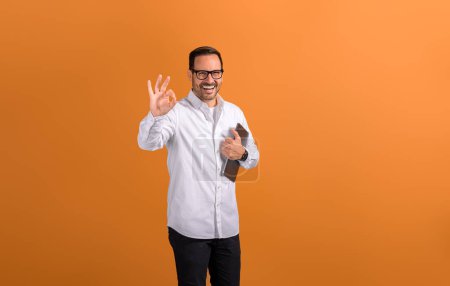 Retrato de hombre de negocios feliz con tableta digital que muestra signo OK y posando sobre fondo naranja