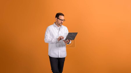 Serious focused businessman in eyeglasses working over laptop while standing on orange background