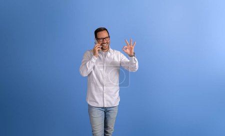 Successful young businessman discussing over phone call and showing OK sign against blue background