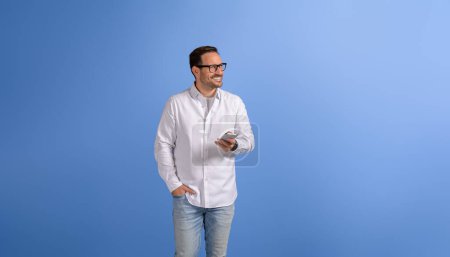 Glücklicher gutaussehender Manager mit Hand in Tasche, Smartphone in der Hand und Blick weg über blauen Hintergrund