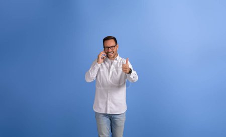 Porträt eines selbstbewussten Geschäftsmannes, der am Telefon telefoniert und auf blauem Hintergrund ein Zeichen mit erhobenem Daumen zeigt