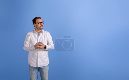 Jeune homme d'affaires réfléchi avec téléphone portable détournant les yeux sérieusement sur fond bleu isolé