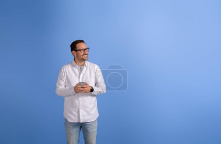 Heureux entrepreneur masculin avec téléphone intelligent regardant loin et pensant idées d'affaires sur fond bleu