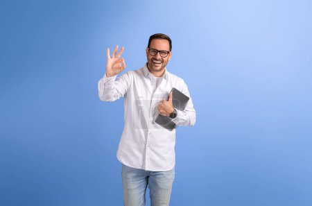 Junger Verkäufer hält digitales Tablet in der Hand und zeigt OK-Zeichen in die Kamera, während er auf blauem Hintergrund posiert