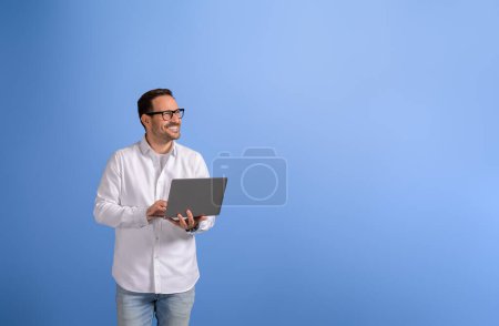 Successful pensive businessman with laptop computer looking away and standing on blue background