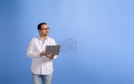 Nachdenklicher männlicher Profi mit drahtlosem Computer, der wegschaut und vor blauem Hintergrund steht