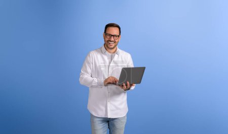 Lächelnder junger Geschäftsmann recherchiert online über Laptop, während er auf isoliertem blauem Hintergrund posiert