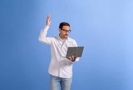 Young businessman raising hand while discussing over video call on laptop against blue background
