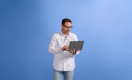 Seriöser gutaussehender Geschäftsmann checkt E-Mails über drahtlosen Computer und steht auf blauem Hintergrund