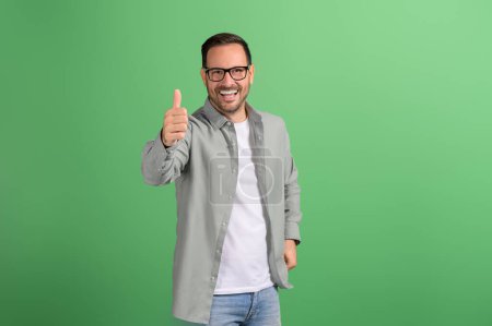 Portrait of positive businessman smiling and showing thumbs up sign on isolated green background