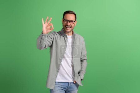 Retrato del guapo freelancer mostrando signo OK y riendo extáticamente sobre fondo verde