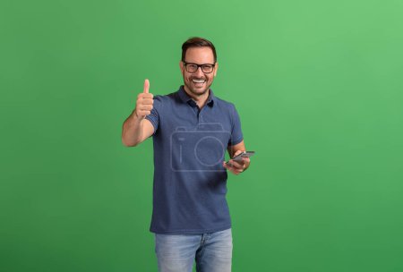 Glücklich selbstbewusster Geschäftsmann, der per Smartphone SMS schreibt und auf grünem Hintergrund ein Zeichen mit Daumen nach oben zeigt