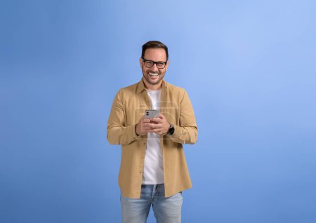 Portrait de joyeux freelance masculin dans les lunettes client de messagerie sur téléphone intelligent sur fond bleu
