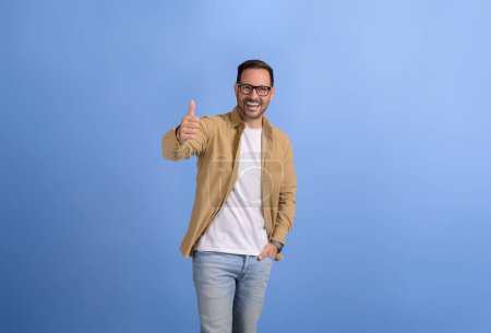 Schöner Unternehmer lächelt ekstatisch und zeigt Daumen hoch, während er vor blauem Hintergrund posiert