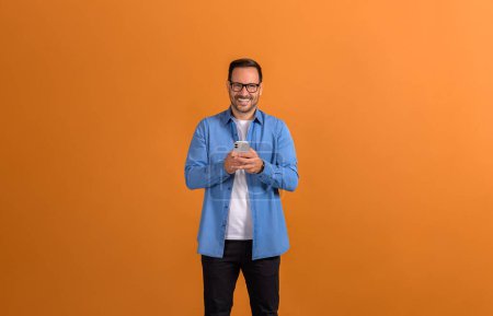 Retrato de feliz joven empresario confiado mensajes de texto a través de teléfono inteligente sobre fondo naranja aislado
