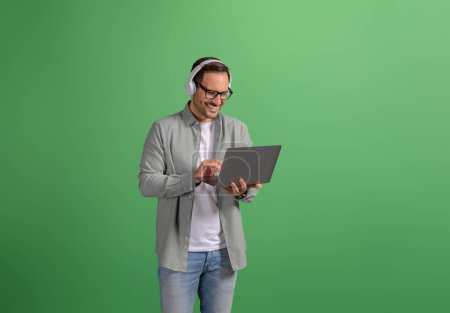 Junger Geschäftsmann trägt Kopfhörer und diskutiert über Videoanruf auf Laptop vor grünem Hintergrund