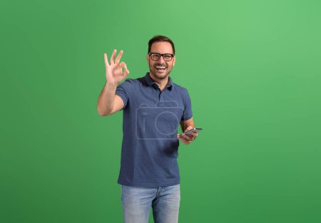 Cheerful handsome entrepreneur messaging over mobile phone and showing OK sign on green background