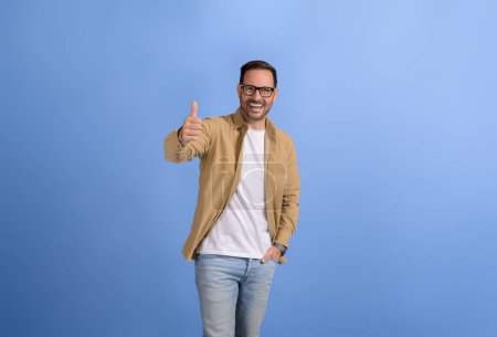 Young businessman with hand in pocket smiling and gesturing thumbs up sign against blue background