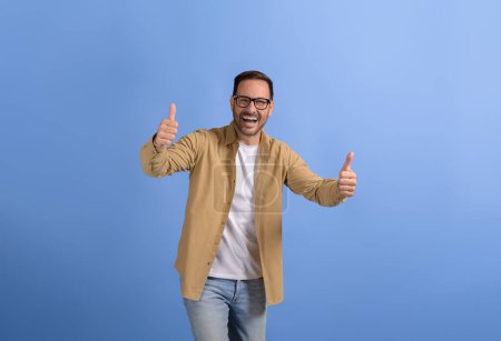 Successful young businessman showing thumbs up signs ecstatically while posing on blue background