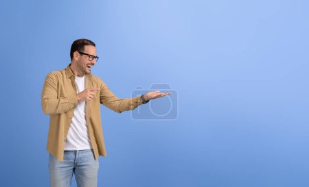 Fröhlicher Verkäufer zeigt auf leere Handfläche und wirbt für neues Produkt auf isoliertem blauem Hintergrund
