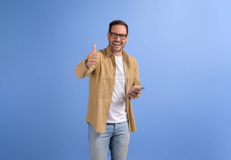 Selbstbewusst glücklich Freelancer SMS über Smartphone und zeigt Daumen nach oben Geste auf blauem Hintergrund
