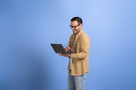 Young businessman smiling and working online over laptop while standing on isolated blue background