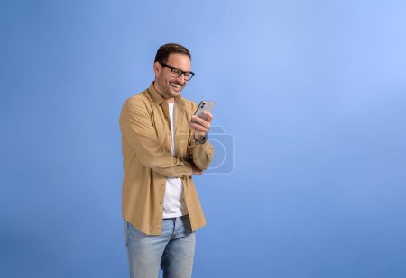 Retrato del gerente guapo sonriente comprobando los mensajes en línea sobre el teléfono móvil sobre fondo azul
